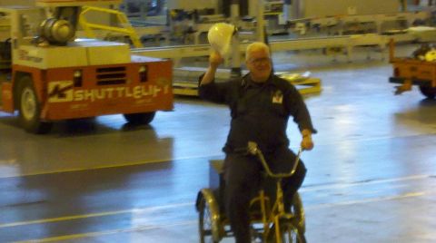 1963 Ford Galaxie - A man waves a hard hat while riding a tricycle in an industrial setting with a shuttle vehicle nearby.