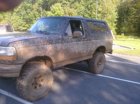 1995 Ford Bronco - This muddy SUV, likely from the early to mid-1990s, is parked on asphalt with trees in the background.