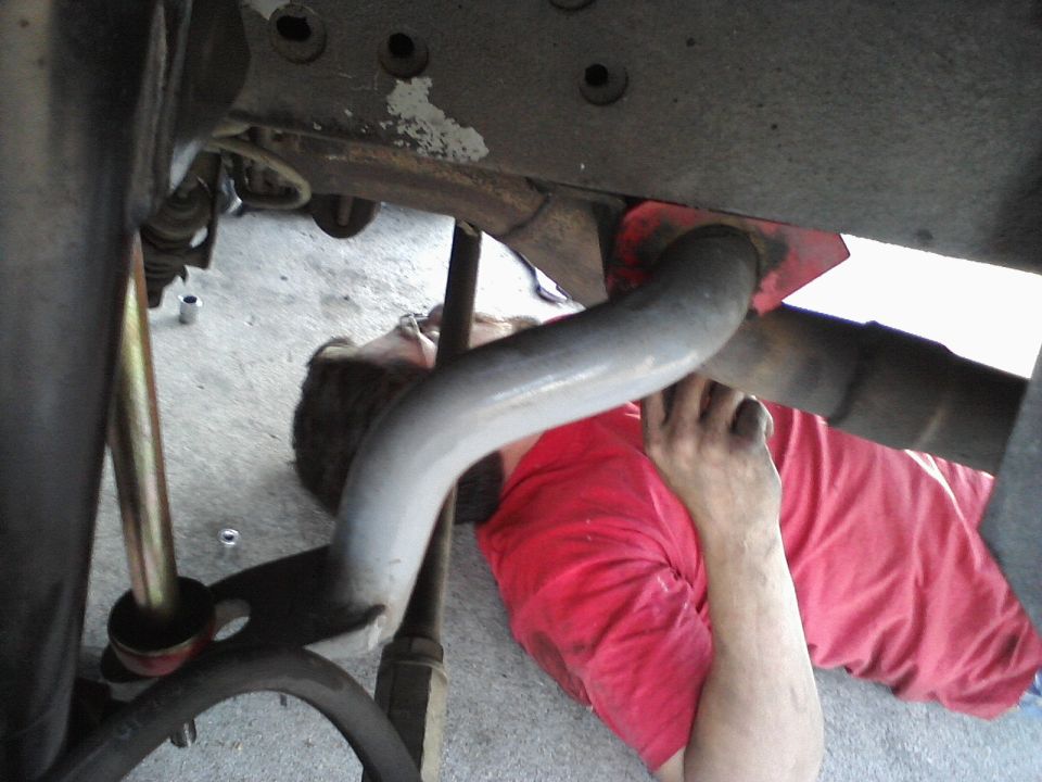 Person in red shirt lying down working on car exhaust pipe and suspension components