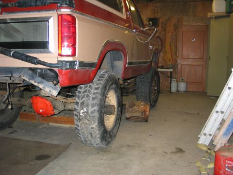1984 Ford Bronco - Rear three-quarter view of a two-tone red and tan lifted Ford pickup or Bronco inside a wood-walled garage, up on jack stands and log block…