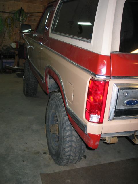 1984 Ford Bronco - Rear driver-side corner of a two-tone red and tan full-size Ford Bronco inside a dim garage, showing the vertical taillight, chrome tailgat…