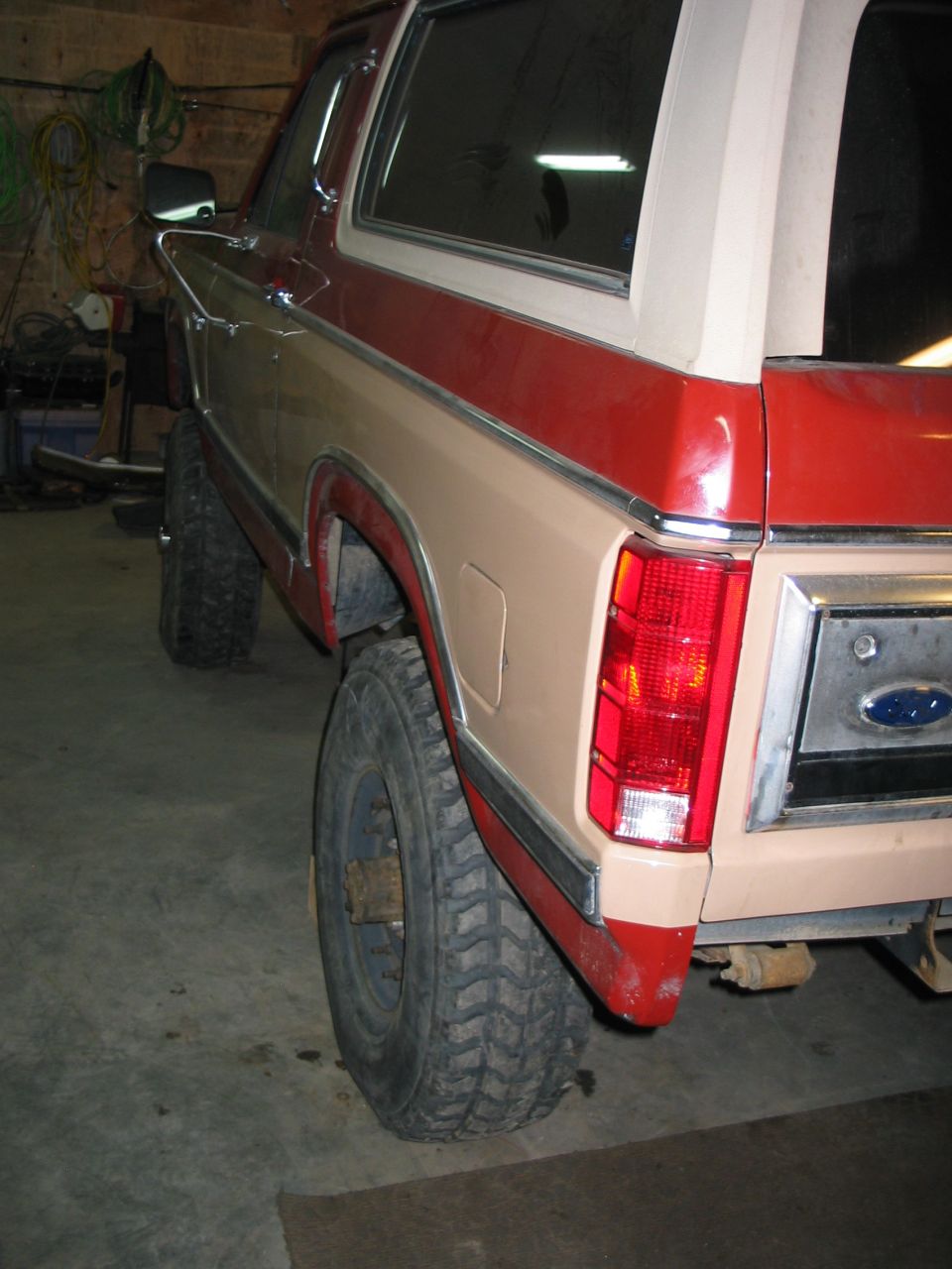 Rear corner of a two-tone red and tan Ford Bronco on mud tires in a garage