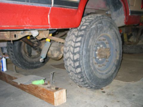1984 Ford Bronco - Low ground-level view alongside a lifted red truck in a garage, showing an axle with aggressive mud-terrain tires on steel wheels, a coil-w…