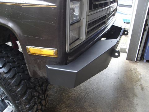 1989 Ford Bronco - Close-up of the front driver-side corner of a dark brown/gray SUV fitted with a gray steel front bumper featuring a D-ring mount tab. The t…
