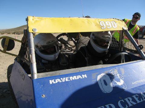 1986 Ford Bronco - Two drivers in helmets sit inside a blue off-road buggy under a yellow sunshade.