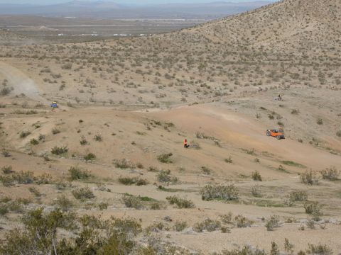 1986 Ford Bronco - Two off-road buggies navigate a dusty desert terrain with sparse vegetation and rolling hills.