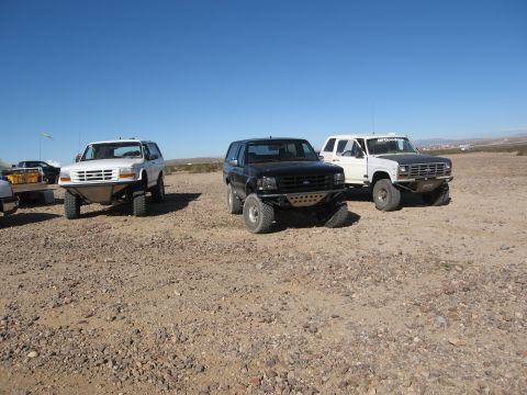 1986 Ford Bronco - Three modified Ford Bronco SUVs are parked in a desert landscape on a sunny day.