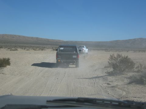 1986 Ford Bronco - Two SUVs travel down a dirt road in a dry, desert landscape under a clear blue sky.