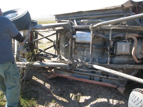 1986 Ford Bronco - The underside of a wrecked vehicle is shown from a low angle, revealing its damaged chassis and mechanical parts.