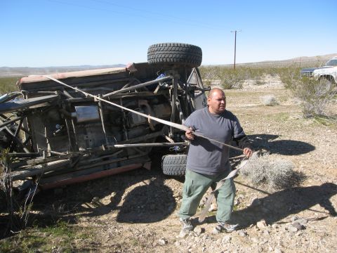 1986 Ford Bronco - A man attempts to right an overturned off-road vehicle in a desert landscape, with another vehicle visible in the distance.