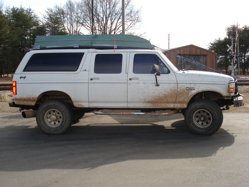 White extended cab pickup truck with a green boat on its roof rack, parked on a road with mud splatters on the side