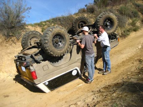 1989 Ford Bronco - An overturned SUV lies on its side on a dirt track, with two individuals observing the scene.