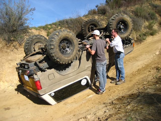 Tan Ford Bronco rolled onto its roof on a dirt trail with two men inspecting it