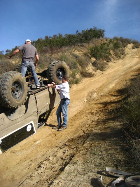 1989 Ford Bronco - Two individuals struggle to right an overturned off-road vehicle on a steep, dusty incline.