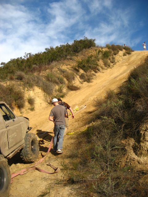 1989 Ford Bronco - Two people are attempting to tow a stuck off-road vehicle up a steep, sandy incline.