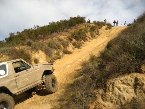 1989 Ford Bronco - This modified SUV is tackling a challenging off-road trail with spectators observing from the ridge above.