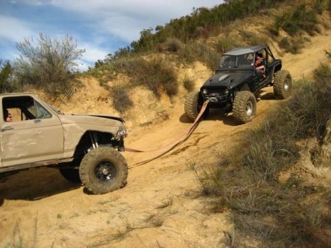 1989 Ford Bronco - Two modified off-road vehicles are engaged in a tow on a steep, sandy incline under a bright sky.