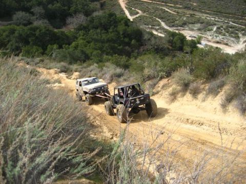 1989 Ford Bronco - Two off-road vehicles navigate a challenging dirt trail in a hilly, arid landscape.
