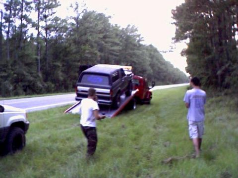 1991 Ford Bronco - A dark SUV is loaded onto a tow truck on the side of a road, with another SUV visible.