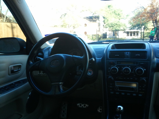 Car interior with black steering wheel, tan door panel, and center console with radio and climate controls. Supermotors.net logo visible