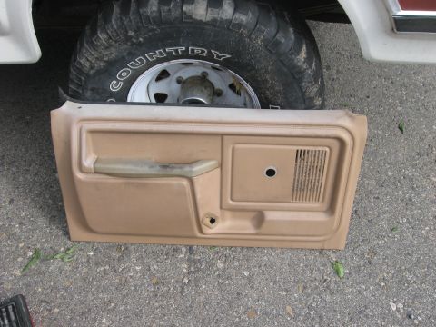 1981 Ford Bronco - A tan door panel is laid out on the ground in front of a vehicle's tire and fender.