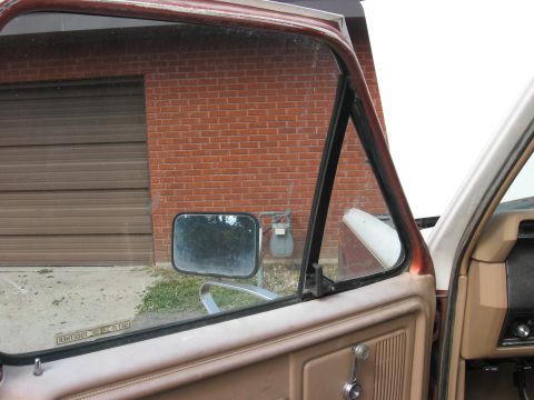 1981 Ford Bronco - View of the interior door panel and wing window, with a brick building and garage door visible outside.