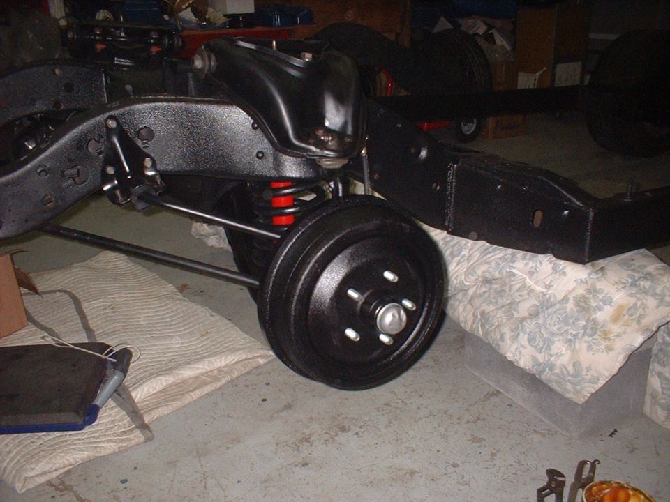 Car chassis frame with suspension components and a drum brake assembly. Red coil springs are visible