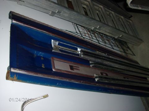 1979 Ford F-150 - A blue Ford tailgate section with chrome trim and an F-O-R-D emblem is shown, with visible rust on its lower edge.