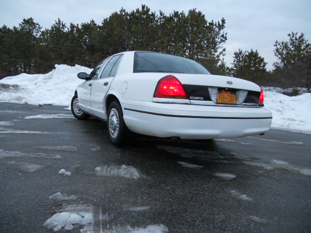 White Ford Crown Victoria sedan parked on wet asphalt with snow piles in background. License plate reads FGV-7260