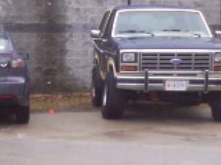 1985 Ford Bronco - A dark blue Ford Bronco is parked next to another vehicle, showing its front grille and headlights.