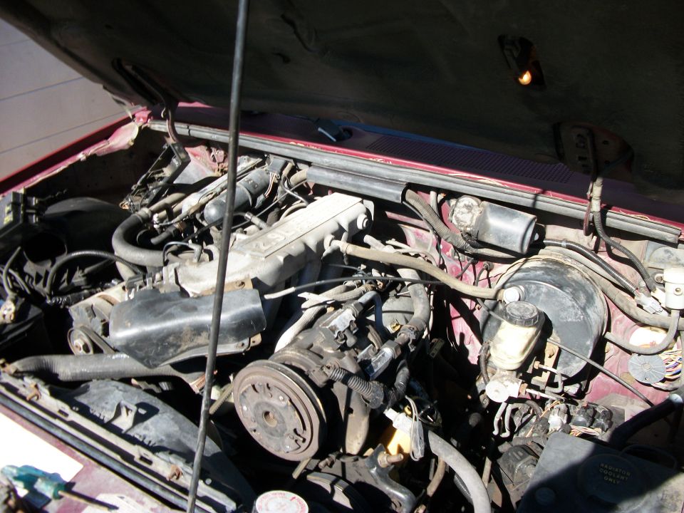 Engine bay of a red truck showing inline engine, valve cover, pulleys and brake booster