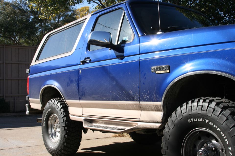 Blue two-door Ford Bronco with tan trim, large mud-terrain tires and side steps, parked outdoors.