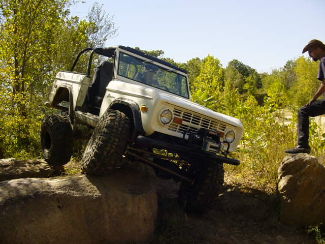 White 1970s Ford Bronco off-roading over rocks, front-left tire elevated