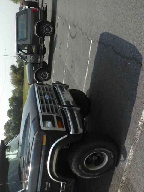 1988 Ford Bronco - Two Ford Broncos are parked in a lot. The front vehicle is dark gray and seen from the front quarter.