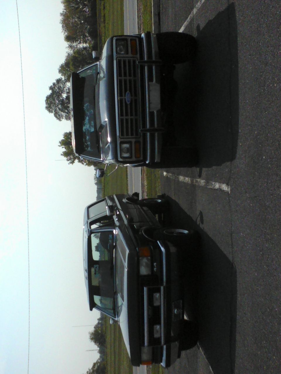 Two dark-colored Ford Broncos parked side-by-side, front view. The front Bronco has a chrome grille and bumper