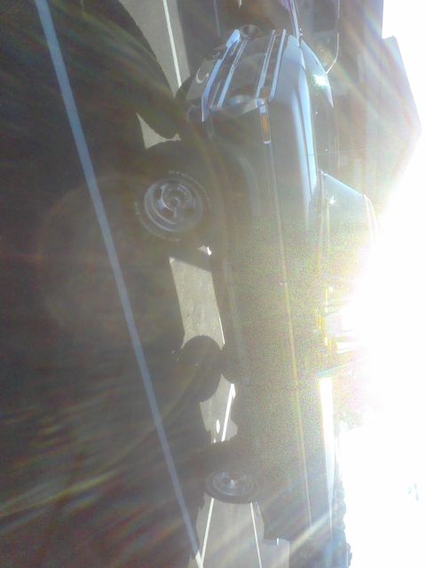 1988 Ford Bronco - A dark SUV with chrome wheels is parked on a street, with bright sunlight glinting off its body.