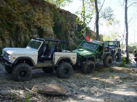 1992 Ford Bronco - Several modified off-road vehicles are parked on a rocky trail, with a steep cliff face in the background.
