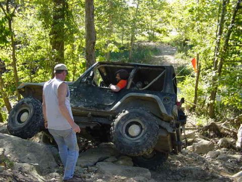 1992 Ford Bronco - A person stands next to a modified off-road vehicle navigating a challenging rocky terrain.