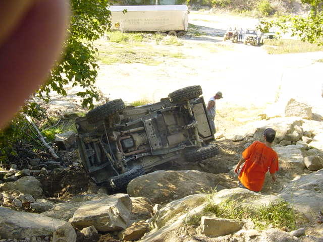 Off-road vehicle overturned on rocks, with two people nearby. Another vehicle and trailer are visible in the background