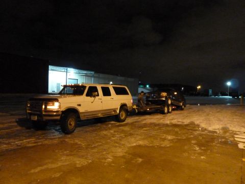 1993 Ford C-150 (Centurion Conversions) - White Ford SUV towing a car on a trailer at night in a parking lot.