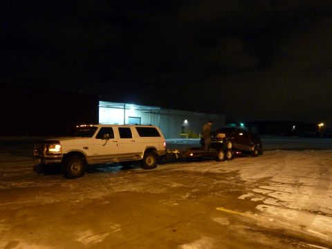 1993 Ford C-150 (Centurion Conversions) - A white Ford F-Series SUV tows a car on a trailer in a snowy lot at night.