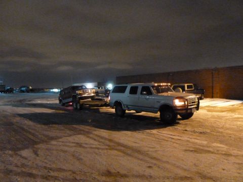1993 Ford C-150 (Centurion Conversions) - A white Ford SUV tows a dark SUV on a trailer in a snowy lot at night.
