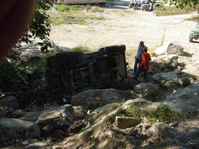 Off-road vehicle flipped on its side in rocky terrain with two people nearby