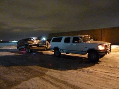 1993 Ford C-150 (Centurion Conversions) - A white Ford SUV tows a dark SUV on a trailer in a snowy lot at night.