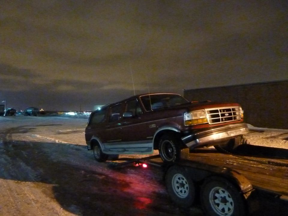 Maroon two-tone Ford Bronco loading onto a tandem-axle flatbed trailer at night in snow