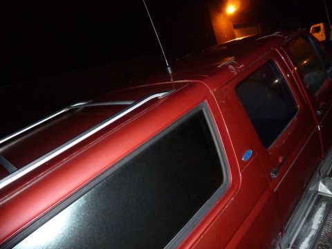1993 Ford C-150 (Centurion Conversions) - The side of a red SUV is visible, with a roof rack and tinted windows. The vehicle appears to be in a dark outdoor setting.
