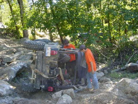 1992 Ford Bronco - An off-road vehicle has overturned on a rocky trail, with two individuals observing the scene.