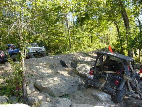 1992 Ford Bronco - Multiple off-road vehicles are seen traversing a challenging rocky path in a wooded area.