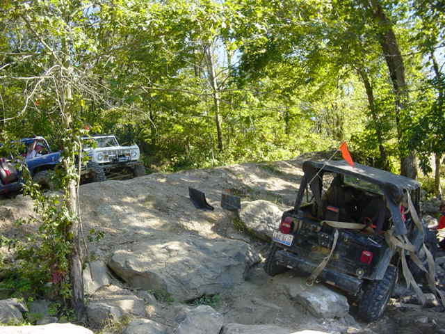 Several off-road vehicles navigating a rocky trail with trees in the background. One vehicle has an orange flag