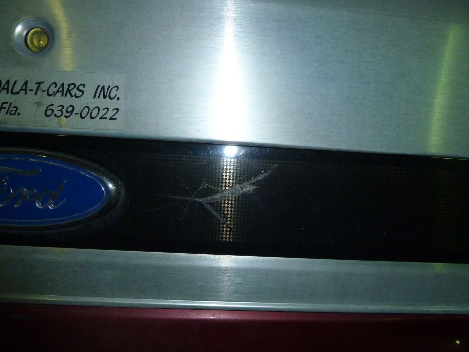 Close-up of a Ford emblem on a vehicle's rear panel. A sticker for 'PALA-T-CARS INC.' with a phone number is visible above it